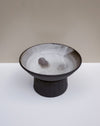 A handmade ceramic pedestal bowl with a dark charcoal exterior and a textured, speckled off-white interior, set against a minimalist beige and white background.  Scree Ceramics | Fodder | Hope Collection