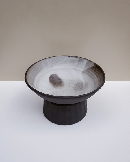 A handmade ceramic pedestal bowl with a dark charcoal exterior and a textured, speckled off-white interior, set against a minimalist beige and white background.  Scree Ceramics | Fodder | Hope Collection