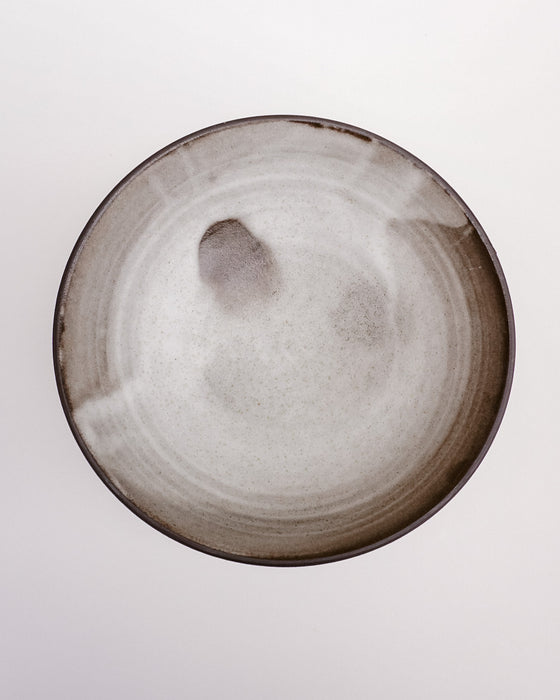 A round ceramic plate with a matte, speckled finish, featuring a dark brown rim and subtle hand-painted gray and brown accents on the interior, shown against a plain white background. Scree Ceramics | Fodder | Hope Collection