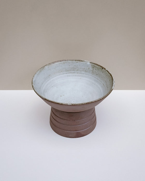 A handmade ceramic pedestal bowl with a textured, earthy brown base and a light gray glazed interior, set against a neutral beige and white background. Scree Ceramics | Fodder | Hope Collection