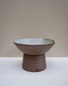 A handmade ceramic pedestal bowl with a textured brown exterior and a light gray glazed interior, set against a neutral beige and white background. Scree Ceramics | Fodder | Hope Collection
