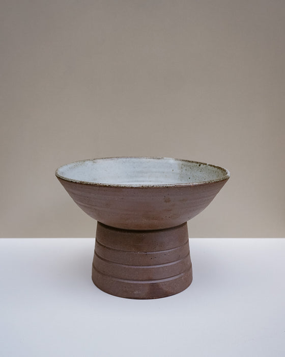 A handmade ceramic pedestal bowl with a textured brown exterior and a light gray glazed interior, set against a neutral beige and white background. Scree Ceramics | Fodder | Hope Collection