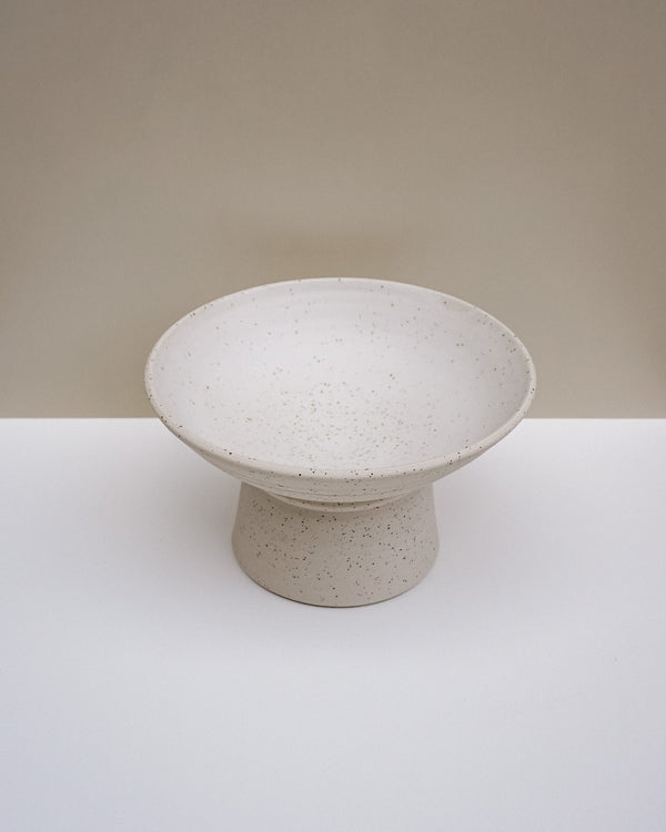 A speckled ceramic pedestal bowl in a warm off-white or cream tone, set against a minimalist beige and white studio background. Scree Ceramics | Fodder | Hope Collection