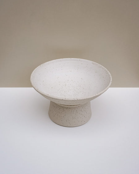 A speckled ceramic pedestal bowl in a warm off-white or cream tone, set against a minimalist beige and white studio background. Scree Ceramics | Fodder | Hope Collection