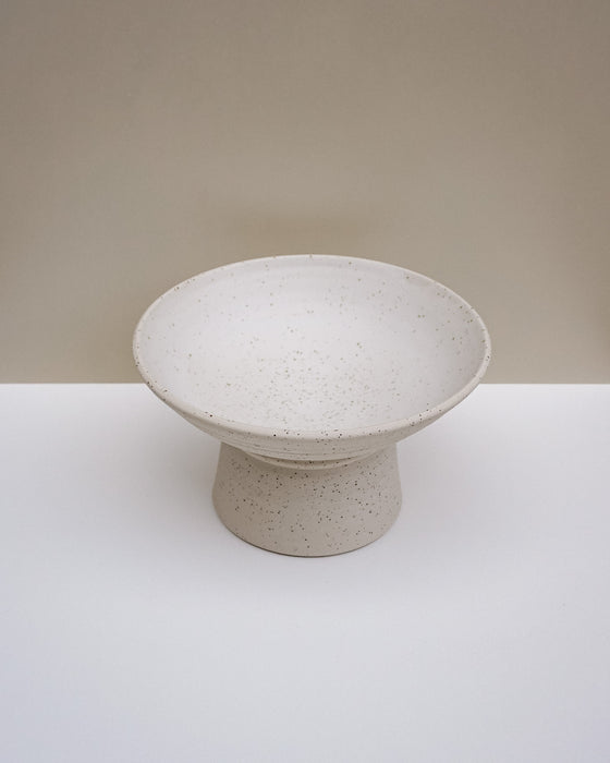 A speckled ceramic pedestal bowl in a warm off-white or cream tone, set against a minimalist beige and white studio background. Scree Ceramics | Fodder | Hope Collection