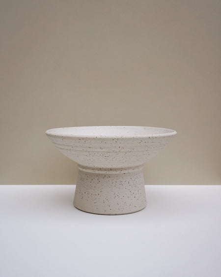 A speckled ceramic pedestal bowl in a warm off-white glaze, set against a minimalist beige and white studio background. Scree Ceramics | Fodder | Hope Collection