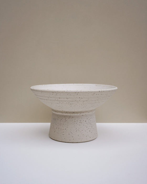 A speckled ceramic pedestal bowl in a warm off-white glaze, set against a minimalist beige and white studio background. Scree Ceramics | Fodder | Hope Collection