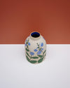 A small ceramic vase with a rounded body, featuring a hand-painted floral pattern of blue flowers and green leaves on a speckled beige glaze. The vase has a dark blue interior and is photographed on a white surface against a terracotta-colored background. Sophie Alda | Hope Collection | Shop Fodder