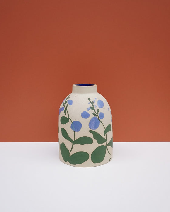 A ceramic vase with a rounded body, featuring a hand-painted blue and green floral pattern on a cream-colored stoneware surface, set against a split orange and white background. Sophie Alda | Hope Collection | Shop Fodder