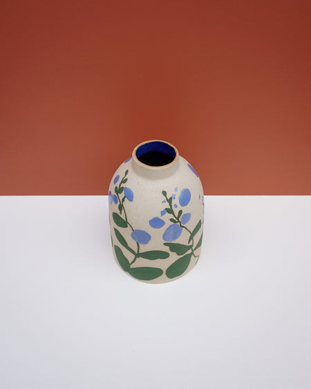 A small ceramic vase with a rounded body, featuring a hand-painted floral pattern of blue flowers and green leaves on a speckled beige glaze. The vase has a dark blue interior and is photographed on a white surface against a terracotta-colored background. Sophie Alda | Hope Collection | Shop Fodder