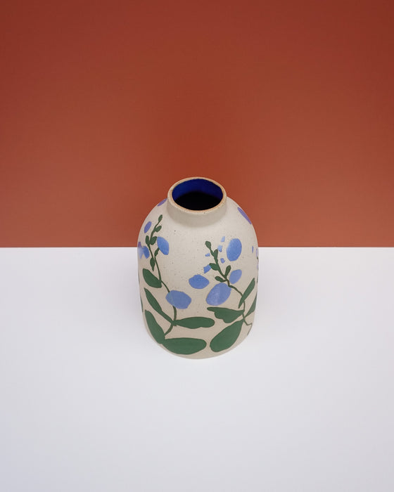 A small ceramic vase with a rounded body, featuring a hand-painted floral pattern of blue flowers and green leaves on a speckled beige glaze. The vase has a dark blue interior and is photographed on a white surface against a terracotta-colored background. Sophie Alda | Hope Collection | Shop Fodder