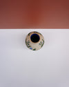 A small ceramic vase with a beige base, decorated with green and blue botanical patterns, featuring a dark blue interior, set against a split white and terracotta background. Sophie Alda | Hope Collection | Shop Fodder