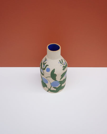 A small ceramic bottle with a beige body, decorated with green leaves and blue berries, featuring a deep blue interior, set against a split white and terracotta background. Sophie Alda | Hope Collection | Shop Fodder