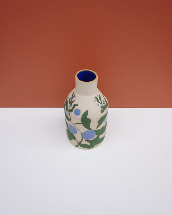 A small ceramic bottle with a beige body, decorated with green leaves and blue berries, featuring a deep blue interior, set against a split white and terracotta background. Sophie Alda | Hope Collection | Shop Fodder