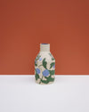 A small ceramic vase with a cream-colored body, decorated with a green and blue floral pattern, standing on a white surface against a terracotta orange background. Sophie Alda | Hope Collection | Shop Fodder
