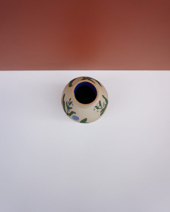 A small ceramic vase with a beige base, decorated with green and blue botanical patterns, featuring a dark blue interior, set against a split white and terracotta background. Sophie Alda | Hope Collection | Shop Fodder