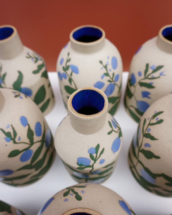 A group of small ceramic bottles with a speckled beige body, decorated with blue flowers and green leaves, featuring a glossy cobalt blue interior glaze, arranged on a white surface against a terracotta background. Sophie Alda | Hope Collection | Shop Fodder