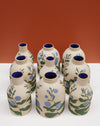 Eight hand-painted ceramic bottles with beige bodies, blue interiors, and green and blue floral motifs, arranged on a white surface against a terracotta background.  Sophie Alda | Hope Collection | Shop Fodder