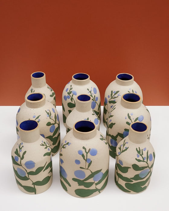 Eight hand-painted ceramic bottles with beige bodies, blue interiors, and green and blue floral motifs, arranged on a white surface against a terracotta background.  Sophie Alda | Hope Collection | Shop Fodder