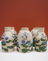 Three handmade ceramic vases with a beige stoneware finish, decorated with a hand-painted pattern of green leaves and blue berries, set against a solid terracotta background. Sophie Alda | Hope Collection | Shop Fodder