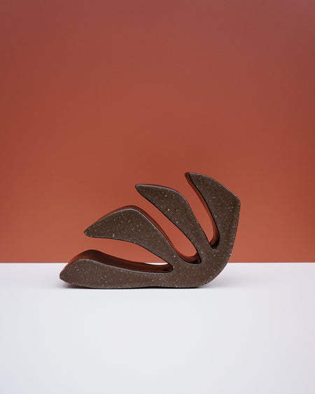 A sculptural, abstract leaf-shaped decorative object with a textured dark brown finish, displayed on a clean white surface against a terracotta-colored background. Studio ERS Ceramics | Hope Collection | Shop Fodder