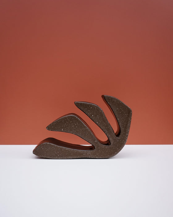 A sculptural, abstract leaf-shaped decorative object with a textured dark brown finish, displayed on a clean white surface against a terracotta-colored background. Studio ERS Ceramics | Hope Collection | Shop Fodder