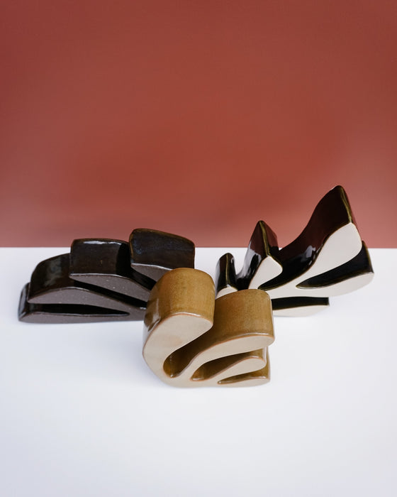 Three abstract sculptural ceramic objects with glossy black and warm beige glazes, arranged on a white surface against a terracotta-colored background. Studio ERS Ceramics | Hope Collection | Shop Fodder