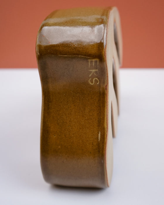 A close-up side view of a glossy ceramic object, likely a candle holder or decorative block, in a warm brown glaze with the letters 'ERS' visible on the side, set against a split white and terracotta background. Studio ERS Ceramics | Hope Collection | Shop Fodder