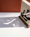 A modern decorative object with a cream-colored rectangular body, black abstract cutout shapes, and a wooden base, photographed on a white surface against a terracotta background with strong shadow play. Studio ERS Ceramics | Hope Collection | Shop Fodder