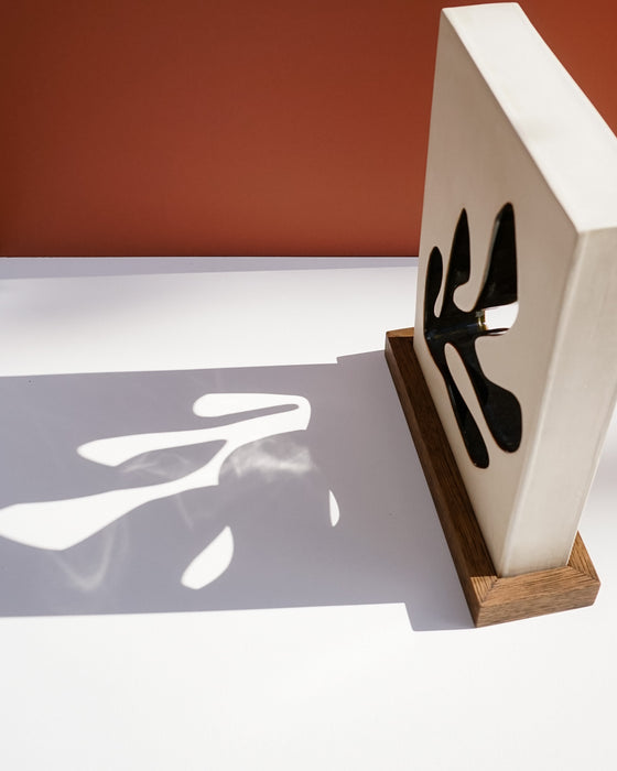 A modern decorative wall art piece featuring a white rectangular panel with black abstract cutout shapes, mounted on a wooden base and casting a bold shadow on a white surface against a terracotta background. Studio ERS Ceramics | Hope Collection | Shop Fodder