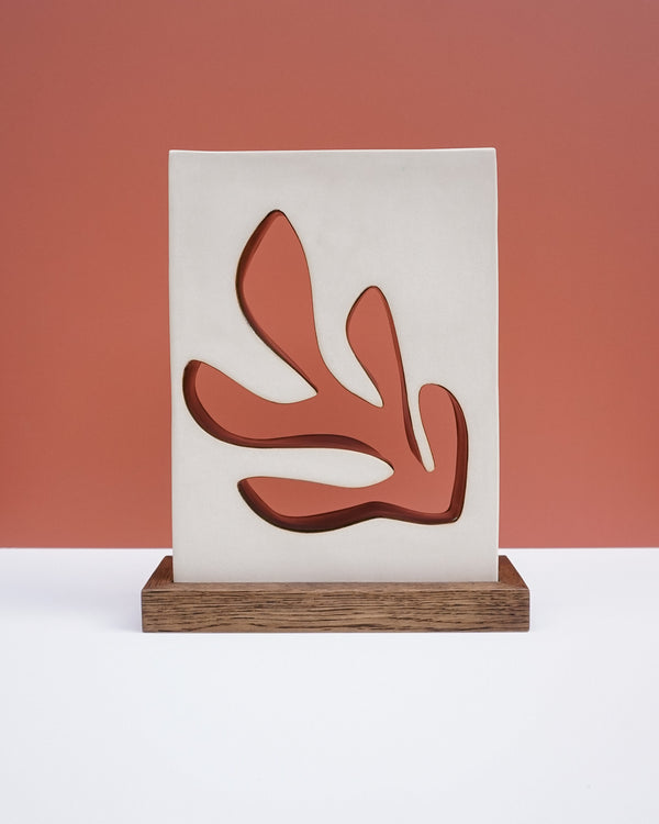 A modern decorative wall plaque or art print featuring an abstract coral-like organic shape in terracotta red, mounted on a vertical off-white panel and displayed on a rectangular wooden base against a terracotta background. Studio ERS Ceramics | Hope Collection | Shop Fodder