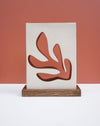 A modern decorative wall plaque or art print featuring an abstract coral-like organic shape in terracotta red, mounted on a vertical off-white panel and displayed on a rectangular wooden base against a terracotta background. Studio ERS Ceramics | Hope Collection | Shop Fodder