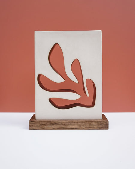 A modern decorative wall plaque or art print featuring an abstract coral-like organic shape in terracotta red, mounted on a vertical off-white panel and displayed on a rectangular wooden base against a terracotta background. Studio ERS Ceramics | Hope Collection | Shop Fodder