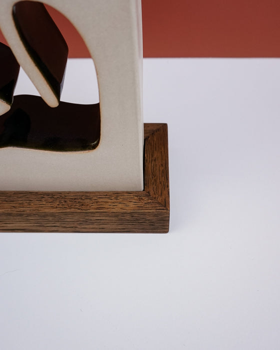 Close-up of a decorative object with a cream-colored upper section, dark brown abstract cutout details, and a medium brown wooden base on a white surface with a terracotta-colored background block. Studio ERS Ceramics | Hope Collection | Shop Fodder