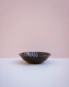 A dark brown ceramic bowl with a glossy finish and gold and white painted stripes, set against a minimalist two-tone background of white and soft pink.