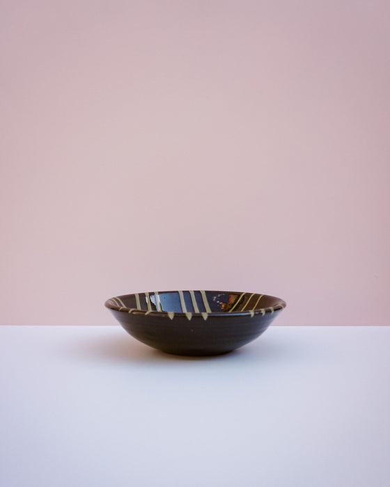 A dark brown ceramic bowl with a glossy finish and gold and white painted stripes, set against a minimalist two-tone background of white and soft pink.