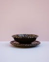 A stack of dark brown ceramic plates with a geometric patterned rim, set against a minimalist two-tone pink and white background.