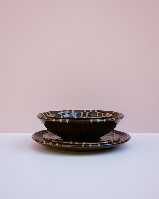 A stack of dark brown ceramic plates with a geometric patterned rim, set against a minimalist two-tone pink and white background.