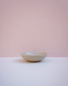 A small ceramic bowl with a textured, speckled off-white glaze, sitting on a white surface against a soft pink background. Lily Pearmain | Fodder