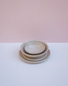 A stack of three nested ceramic plates in a light beige glaze, set against a minimalist background split horizontally into a soft pink upper half and a white lower half.