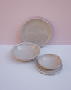 Three handmade ceramic dishes in a light beige glaze, including a large plate, a shallow bowl, and a small plate on a saucer, arranged on a white surface against a pink background. Lily Pearmain Ceramics | Fodder