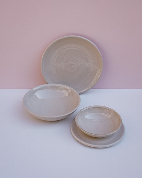 Three handmade ceramic dishes in a light beige glaze, including a large plate, a shallow bowl, and a small plate on a saucer, arranged on a white surface against a pink background. Lily Pearmain Ceramics | Fodder