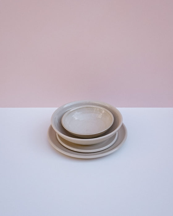A stack of three nested ceramic plates in a light beige glaze, set against a minimalist background split horizontally into a soft pink upper half and a white lower half.