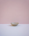 A small ceramic bowl with a speckled off-white and light brown glaze, sitting on a white surface against a solid pink background. Morning Bowl |Lily Pearmain Ceramics | Fodder