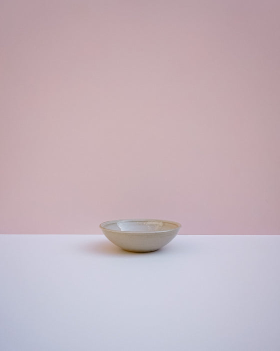 A small ceramic bowl with a speckled off-white and light brown glaze, sitting on a white surface against a solid pink background. Morning Bowl |Lily Pearmain Ceramics | Fodder