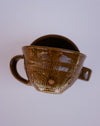 A handmade brown glazed ceramic cup with a textured vertical rib pattern, a side handle, and a small tab on the rim, shown against a plain light gray background.