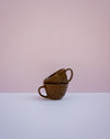 Two stacked brown ceramic cups with a textured, basket-like exterior and looped handles, set against a minimalist two-tone background of soft pink and white.