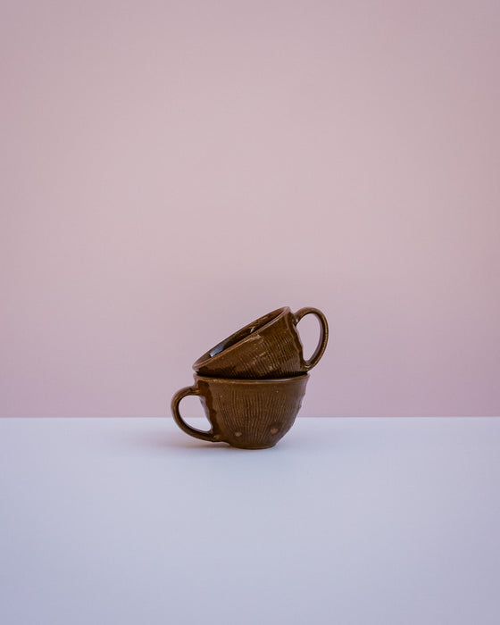 Two stacked brown ceramic cups with a textured, basket-like exterior and looped handles, set against a minimalist two-tone background of soft pink and white.
