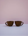Two dark brown glazed ceramic cups with ribbed textures and loop handles, displayed side-by-side on a white surface against a soft pink background.