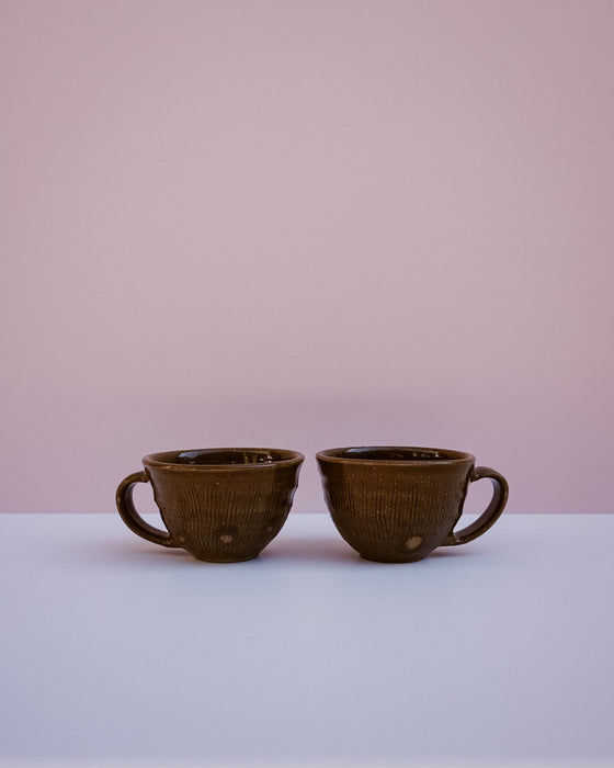 Two dark brown glazed ceramic cups with ribbed textures and loop handles, displayed side-by-side on a white surface against a soft pink background.
